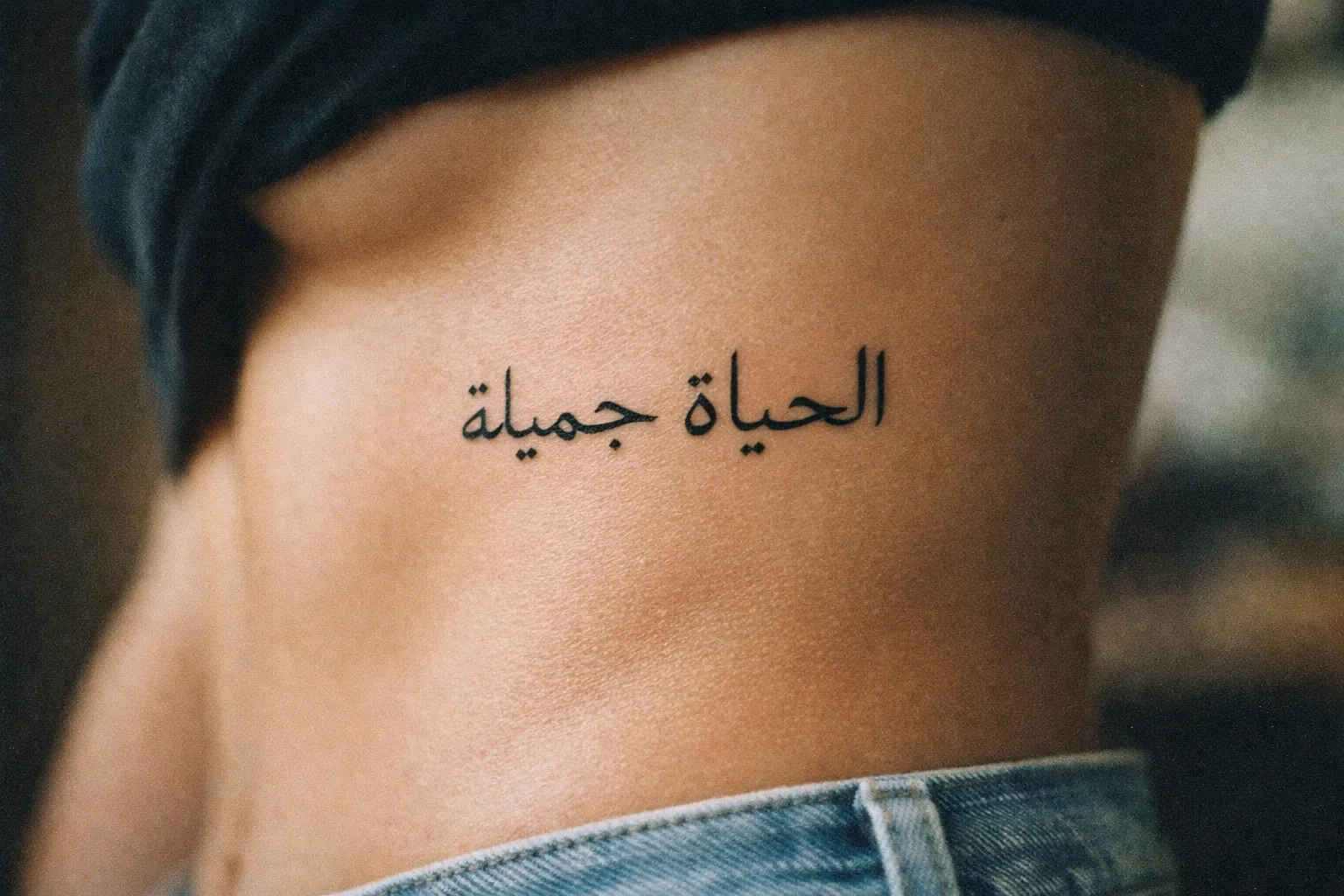 A person's bare side shows an Arabic script tattoo above the waistline of their jeans.