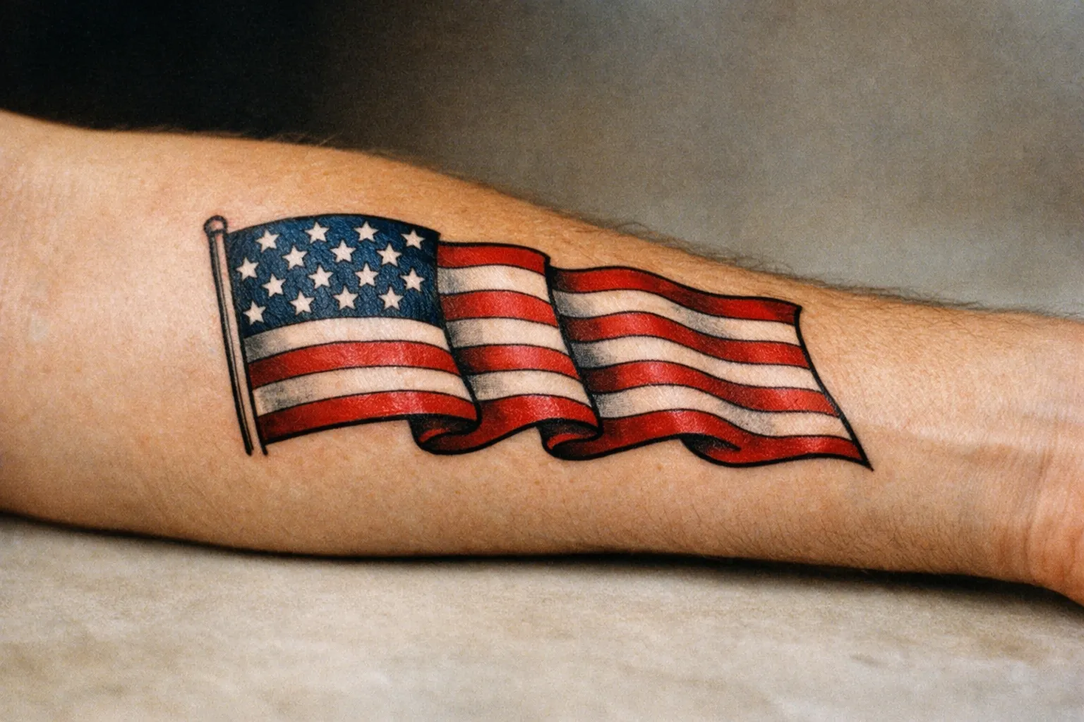 Forearm tattoo of a traditional American flag with bold lines and subtle wear, photographed up close.