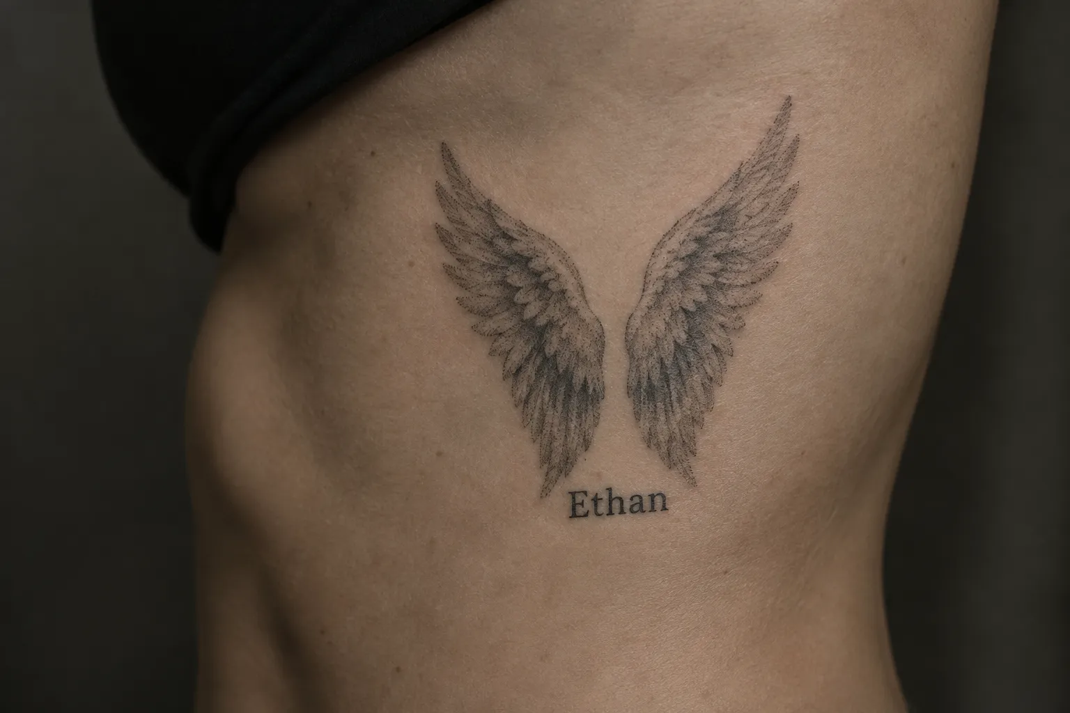 A person's side torso bears an angel wings tattoo with the name Ethan.