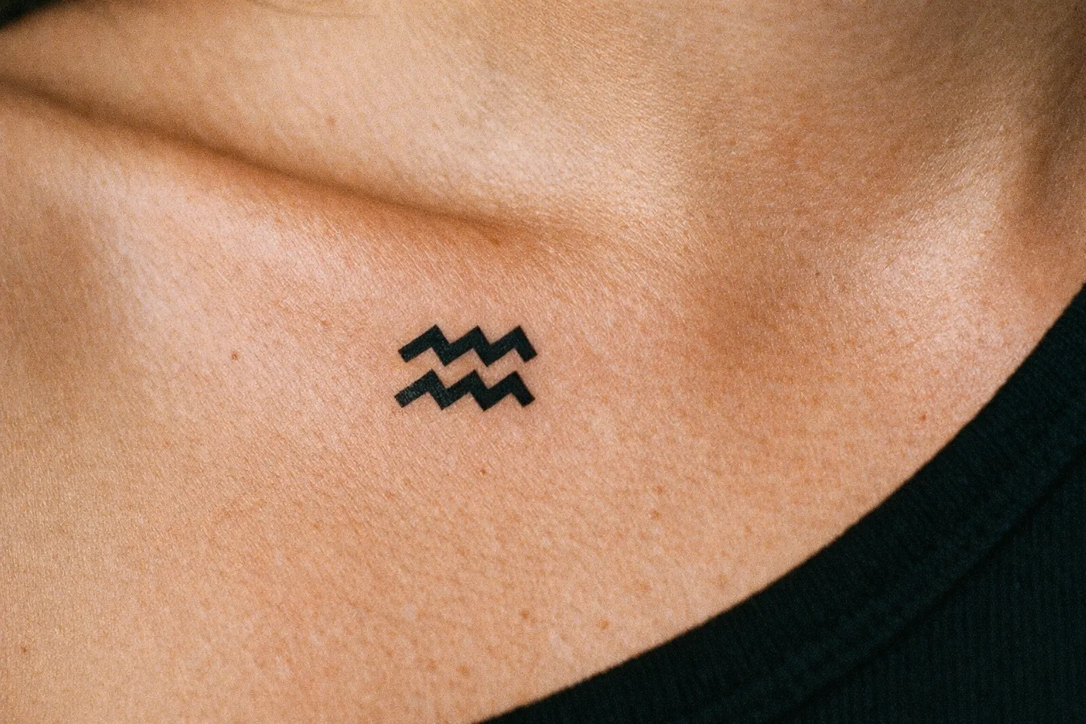 A small Aquarius zodiac symbol tattoo on the upper chest near the collarbone.
