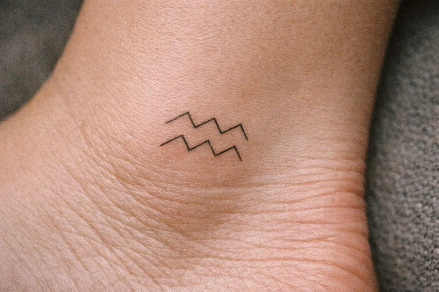 A small Aquarius zodiac symbol tattoo is on the side of a person's ankle.