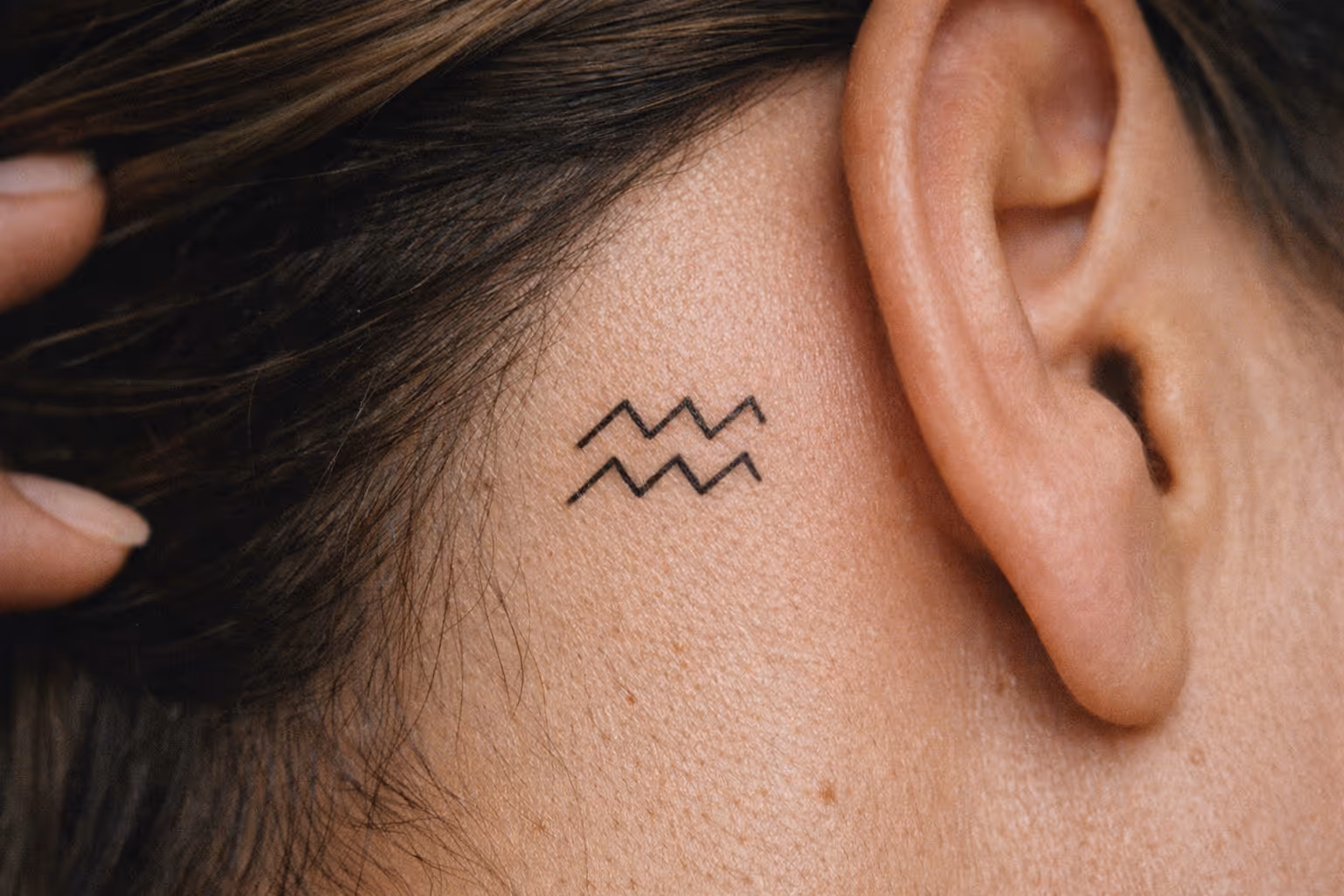 A small Aquarius zodiac symbol tattoo behind a person's ear.