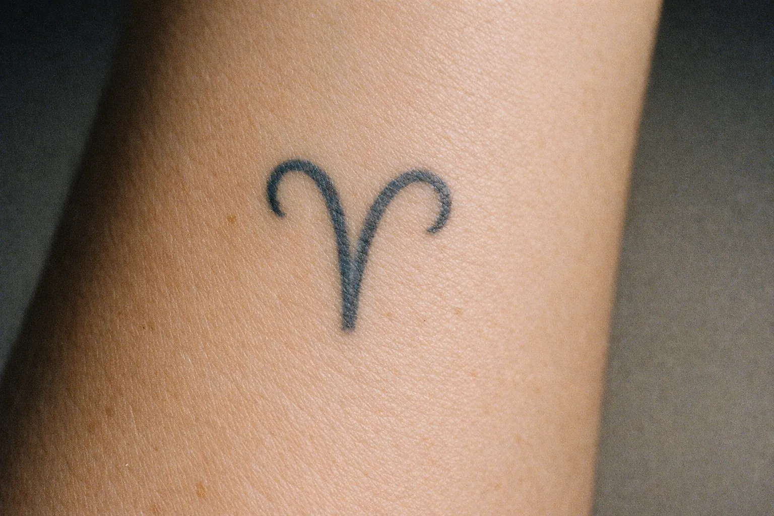Aged Aries glyph tattoo on inner forearm showing slight fading but intact lines