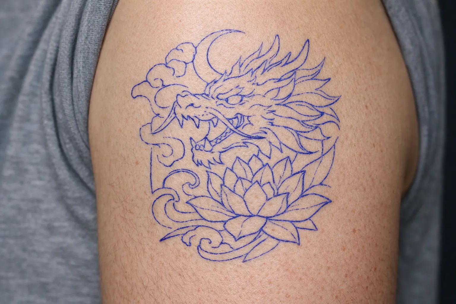 An upper arm with a blue outline tattoo of a dragon and lotus flower.
