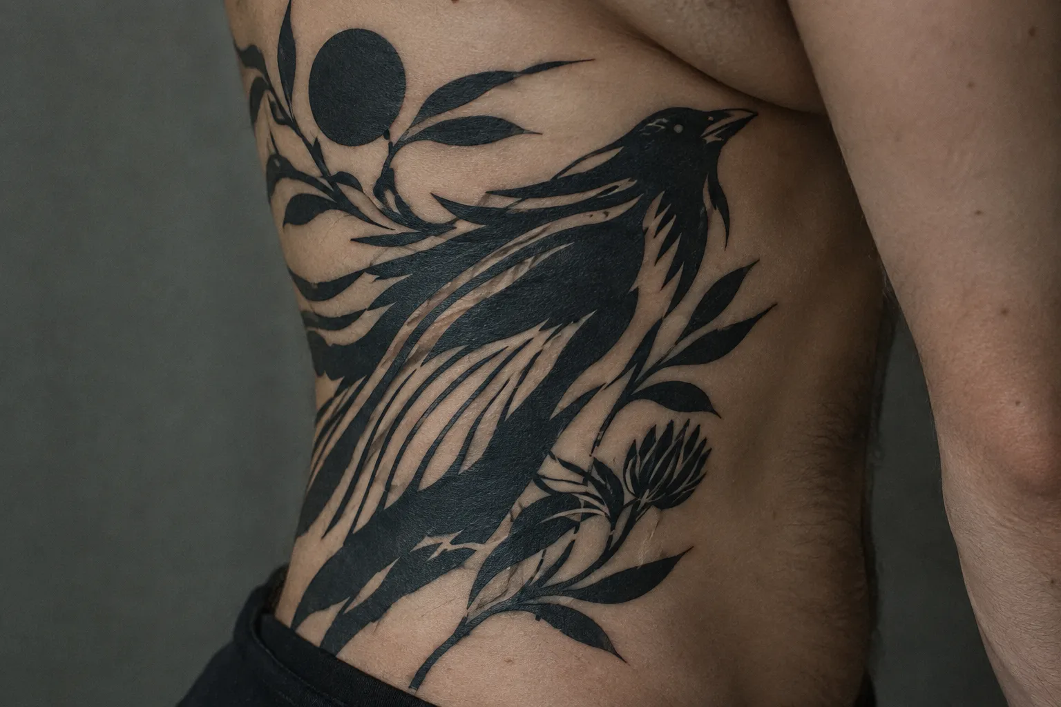 A person's side bears a large blackwork tattoo of a bird and sweeping botanical shapes.
