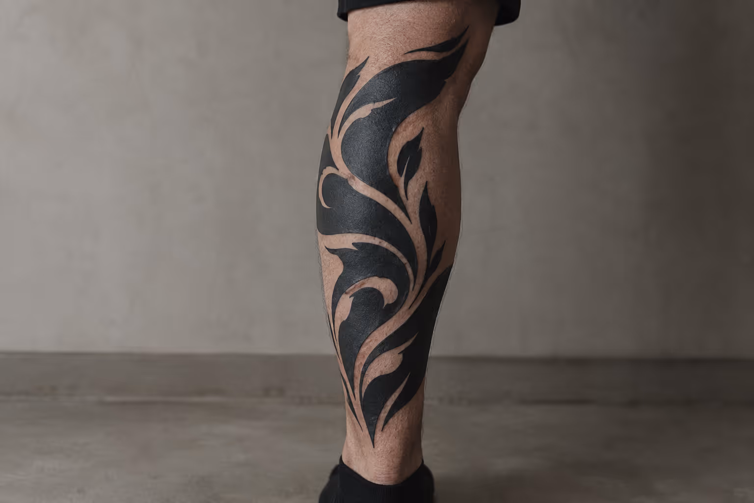 A lower leg with a bold blackwork tattoo stands against a plain gray wall.