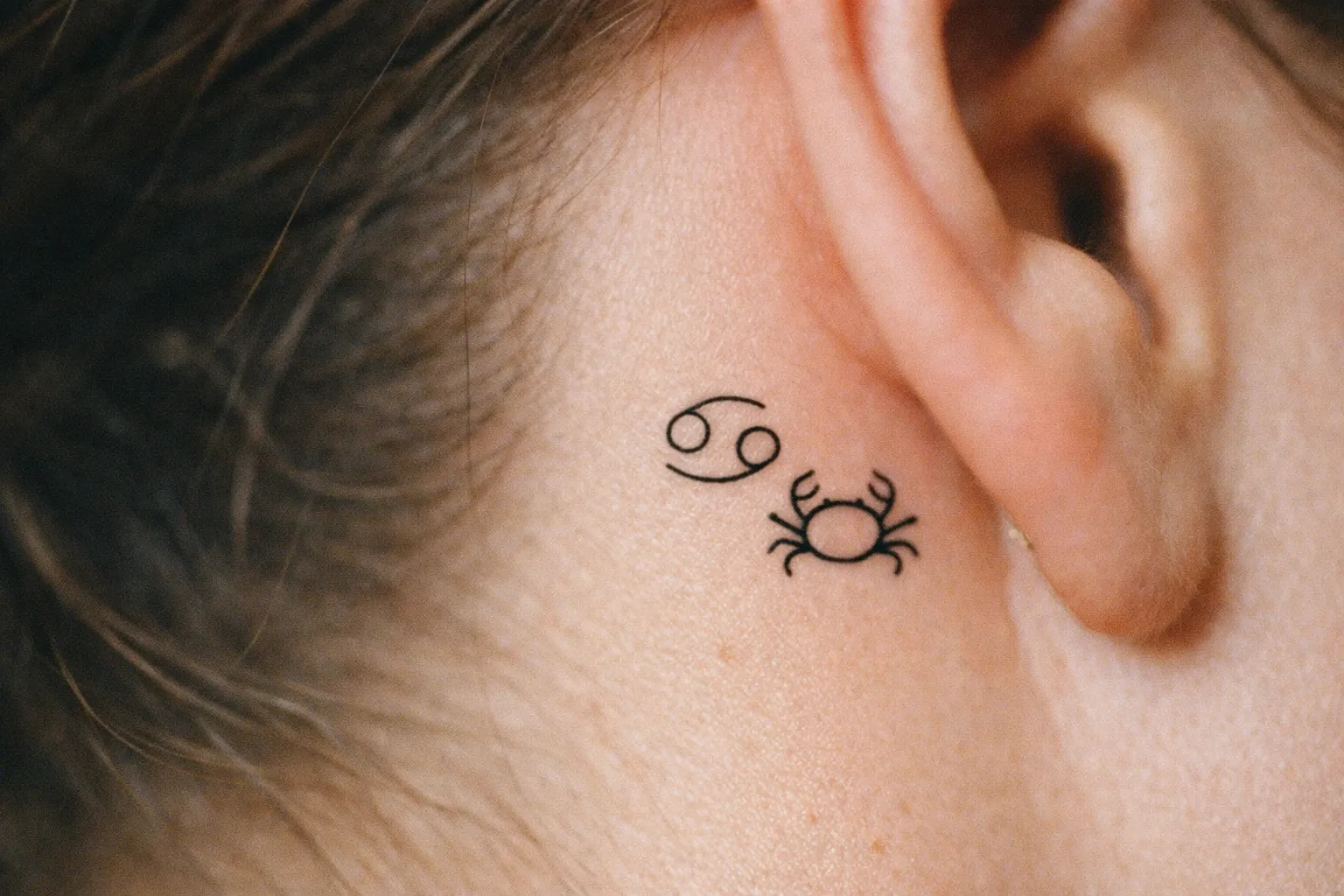 A small crab and Cancer zodiac symbol tattooed behind a person's ear.