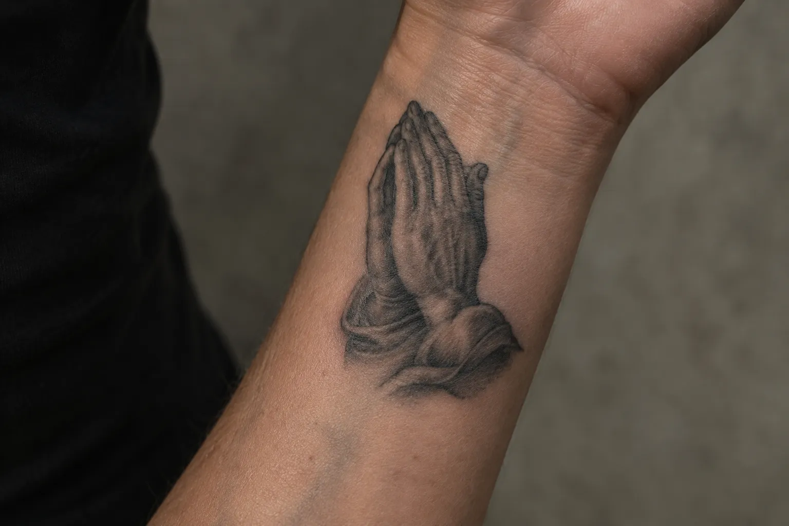 A black-and-gray praying hands tattoo on a person's forearm.