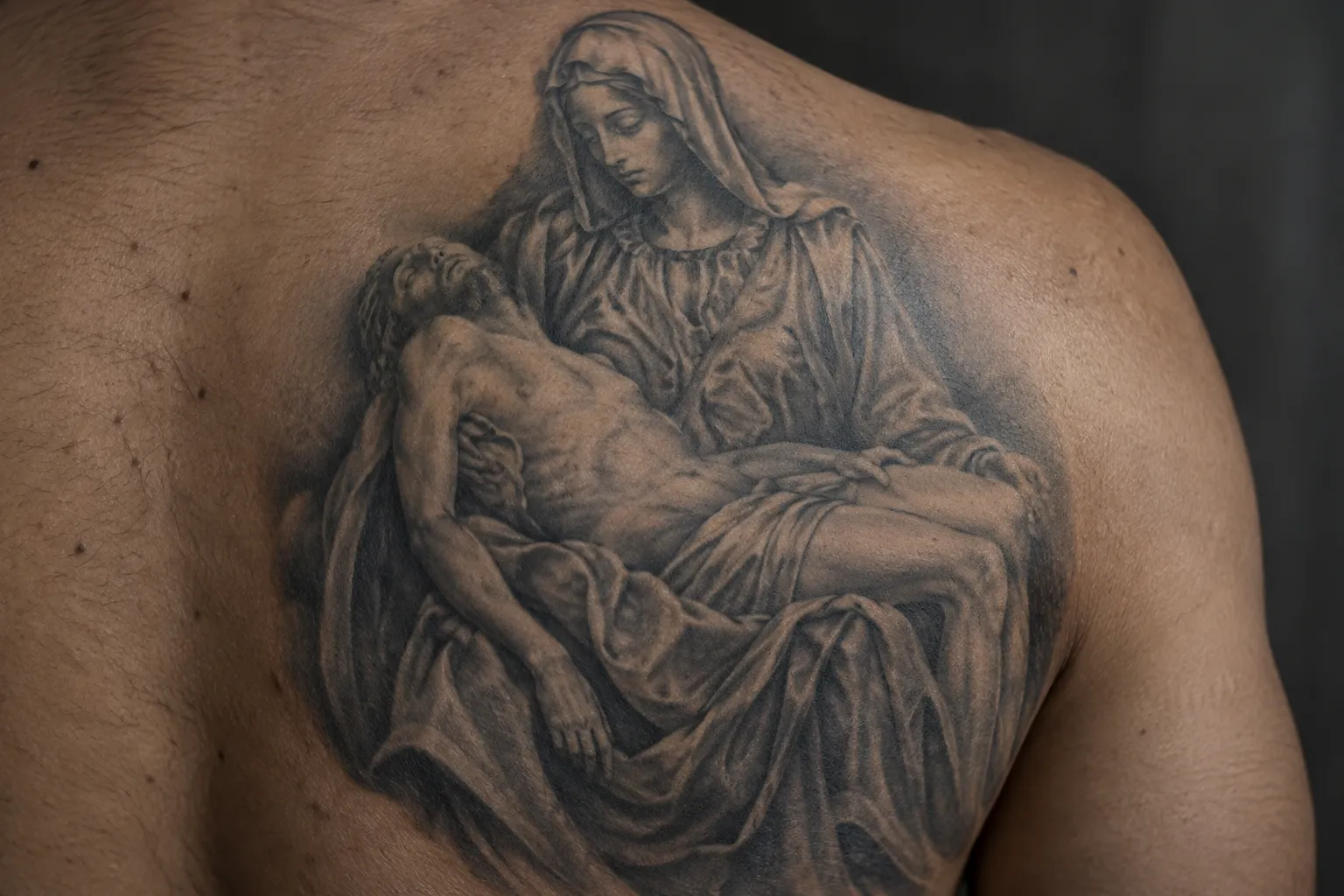 A black-and-grey Pietà tattoo covers a man's upper back and shoulder blade.