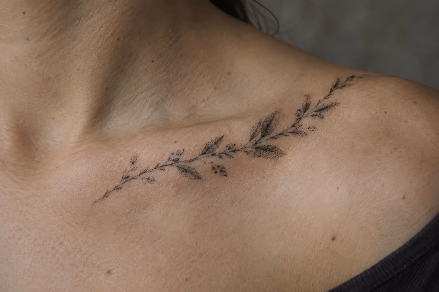 A delicate leafy branch tattoo follows the collarbone on a person's upper chest.