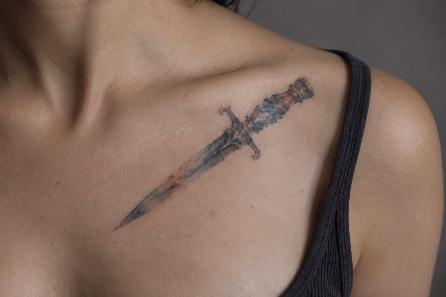 A watercolor dagger tattoo in muted rust and blue sits on a person's upper chest below the collarbone.