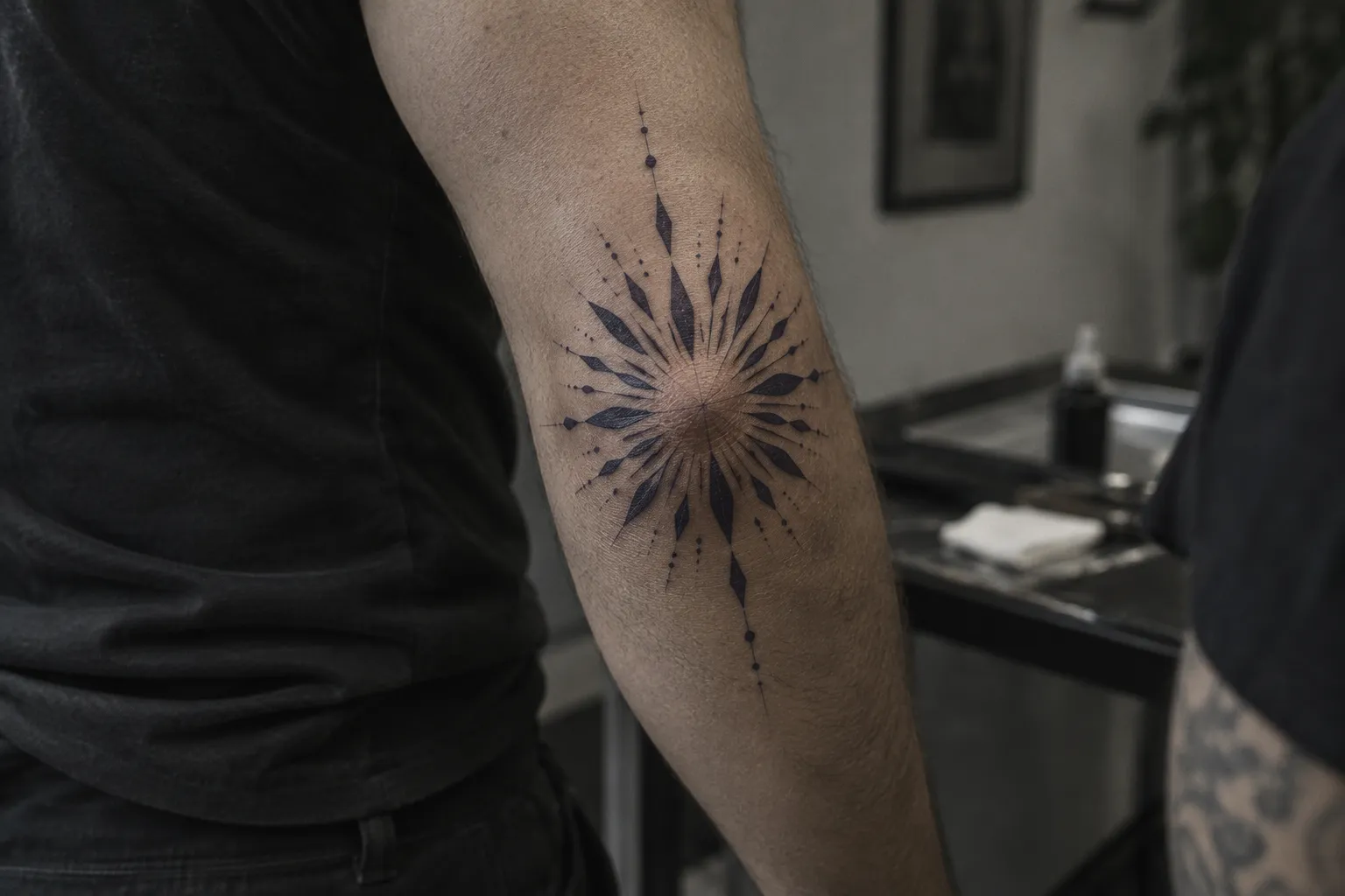 A person shows a geometric sunburst tattoo centered over the elbow in a tattoo studio.
