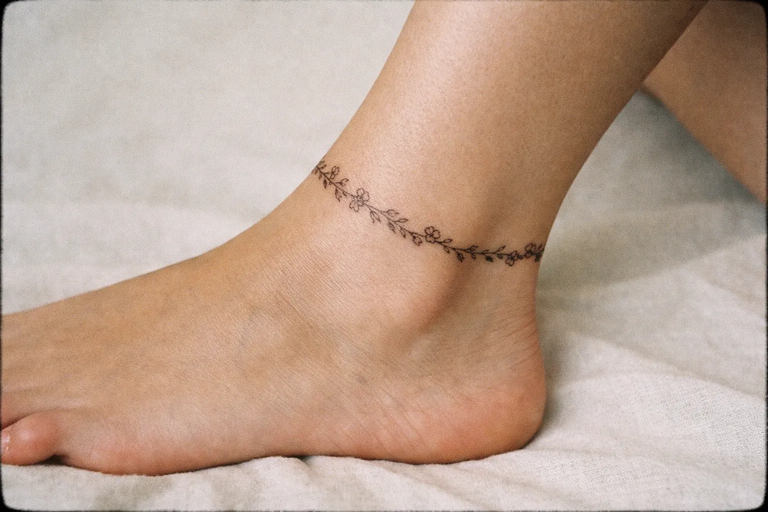 A delicate floral fine line tattoo wraps around an ankle on a bare foot resting on light fabric.