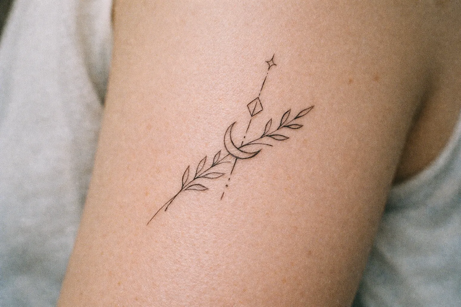 A person's upper arm with a fine line tattoo of a leafy branch, crescent moon, and geometric shapes.