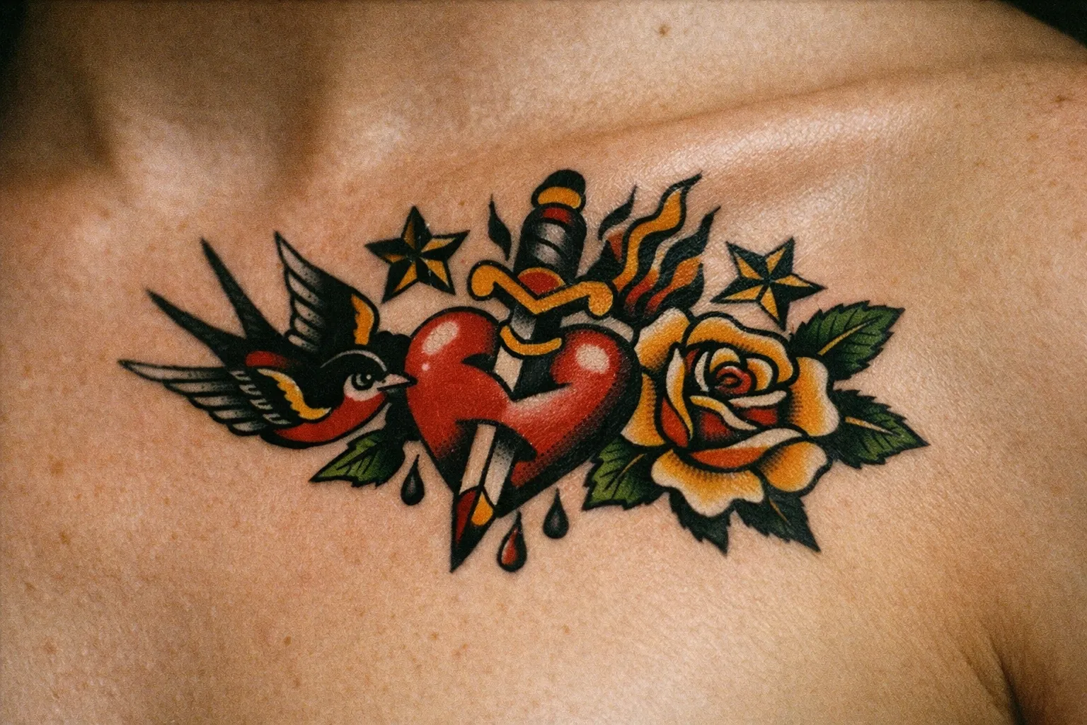 A colorful traditional tattoo of a heart with a dagger, rose, bird, and stars on a person's chest.