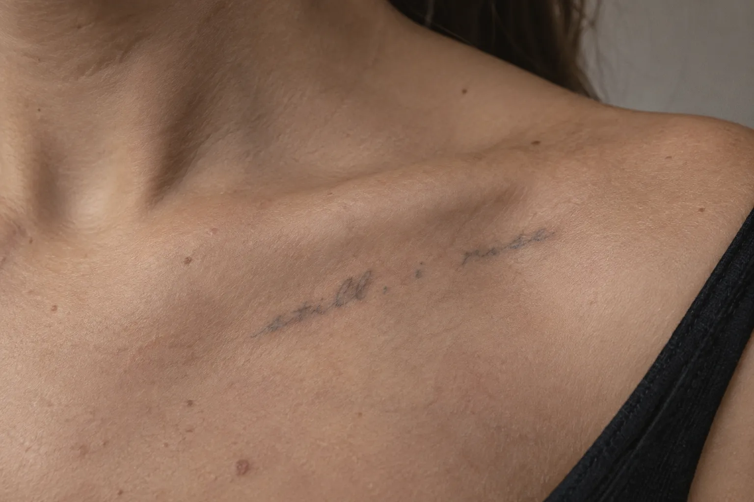 A faint cursive tattoo on a person's upper chest near the collarbone, its thin letters softened and blurred with age.
