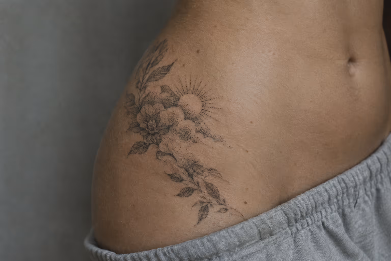 A floral sun tattoo curves across a person's side hip above a gray waistband.