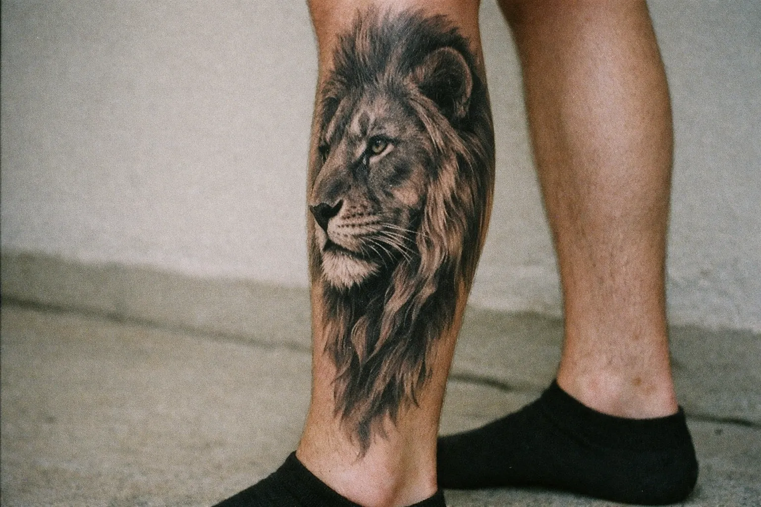 A person with a realistic lion head tattoo on their lower leg stands on a concrete surface.