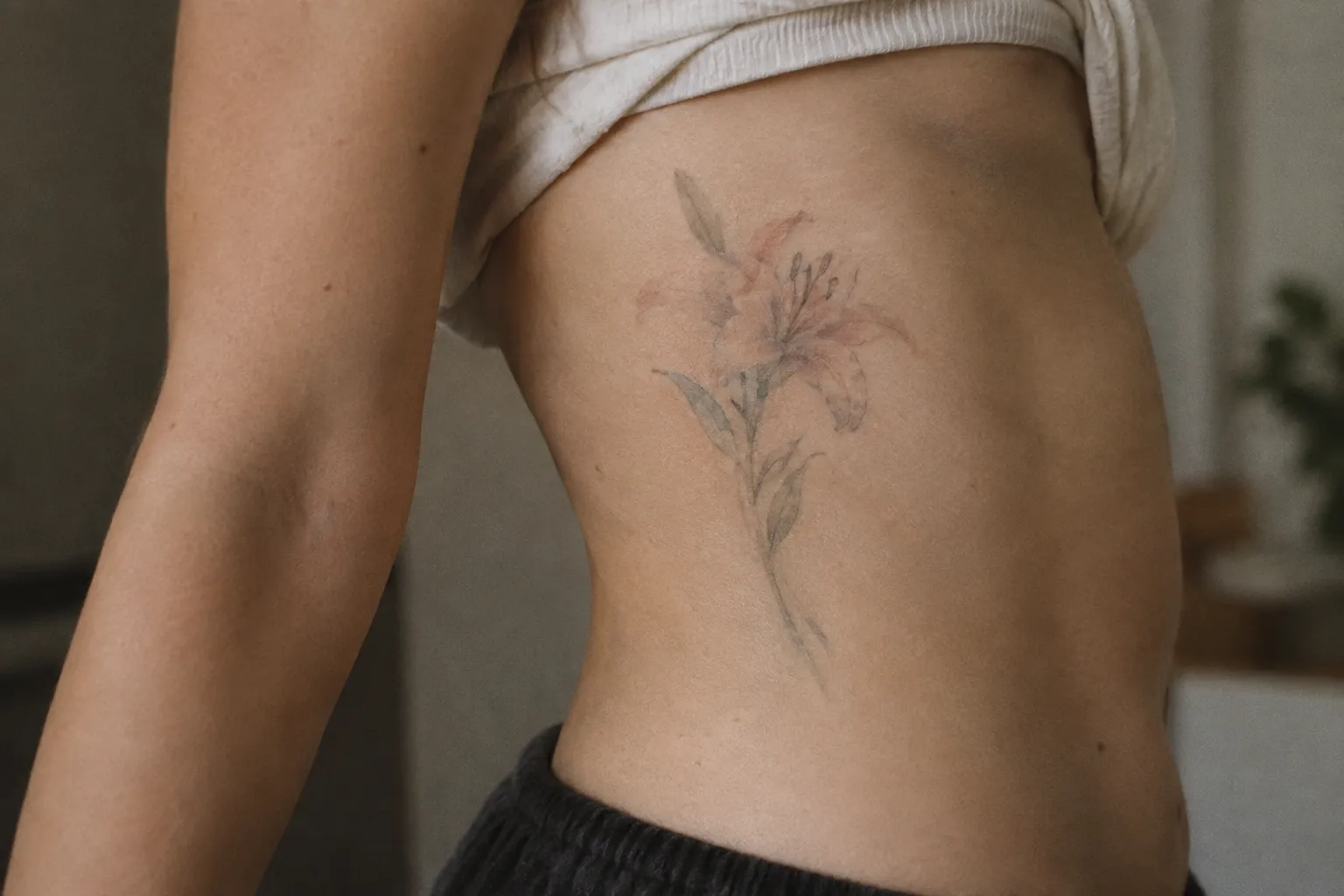A person has a faded pink lily tattoo on their side in an indoor setting.