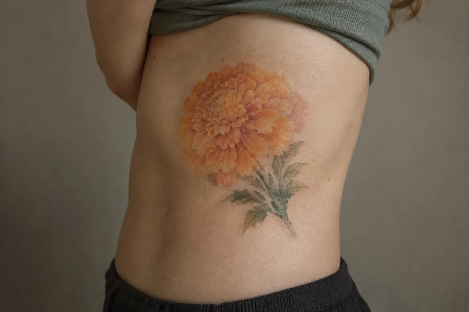 A soft watercolor marigold tattoo on a person's ribs, its orange petals drawn without a dark outline.