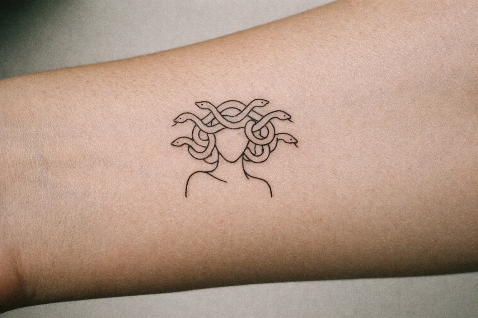 A minimalist Medusa tattoo on the inner forearm, using a single line to form a snake motif without facial features.