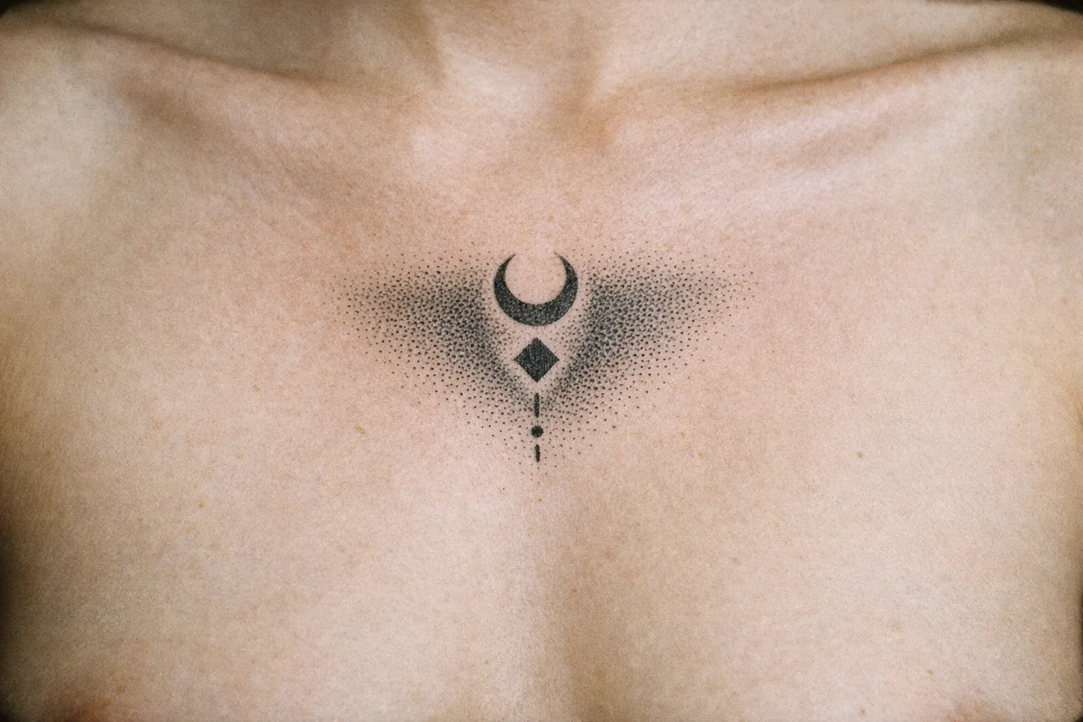 A geometric black ink tattoo is centered on a person's upper chest below the collarbones.
