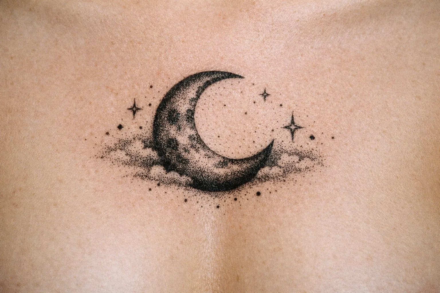 A crescent moon tattoo with clouds and stars is inked on a person's upper chest.