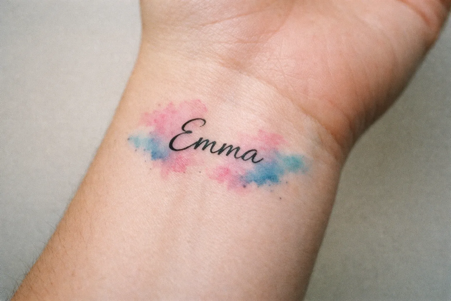 A wrist with a tattoo of the name 'Emma' over a pink and blue watercolor background.