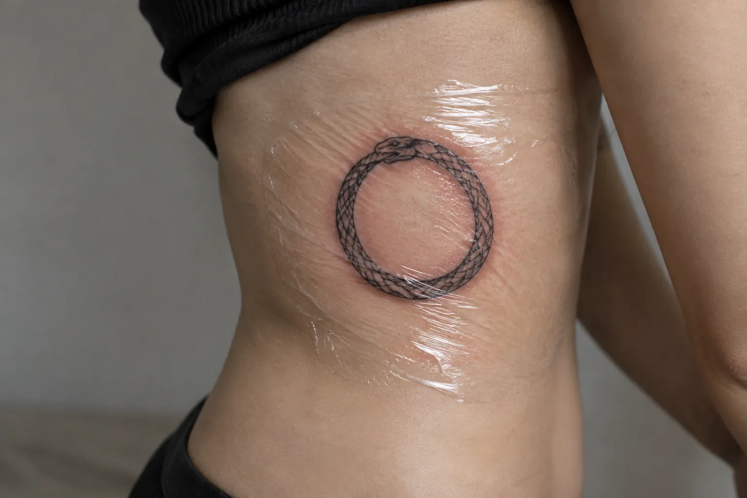A person's side shows a circular ouroboros snake tattoo beneath clear protective film.
