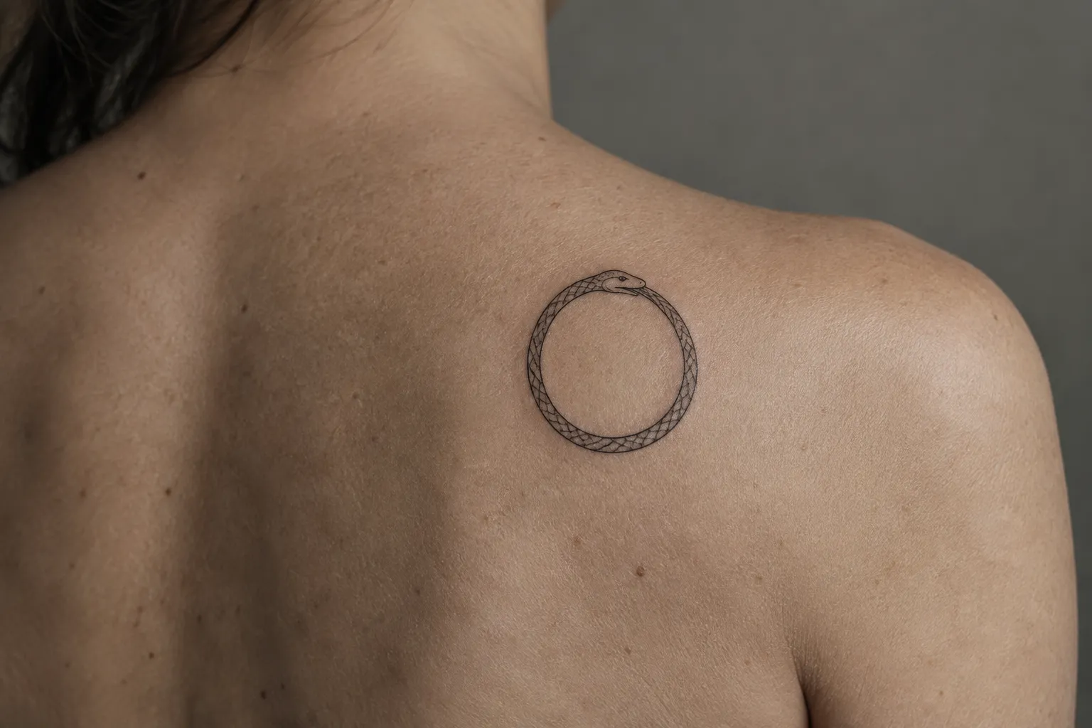 A small fine-line ouroboros near a person's shoulder blade, its head-to-tail join clearly readable.