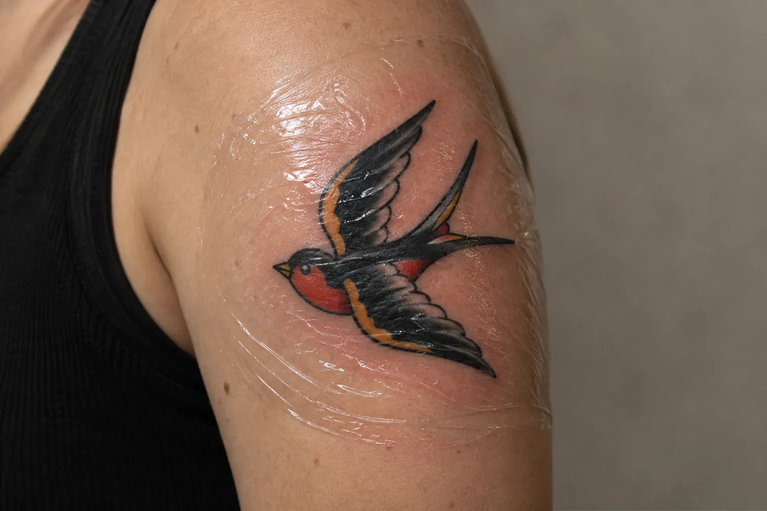 A swallow tattoo on a person's upper arm beneath clear protective film.