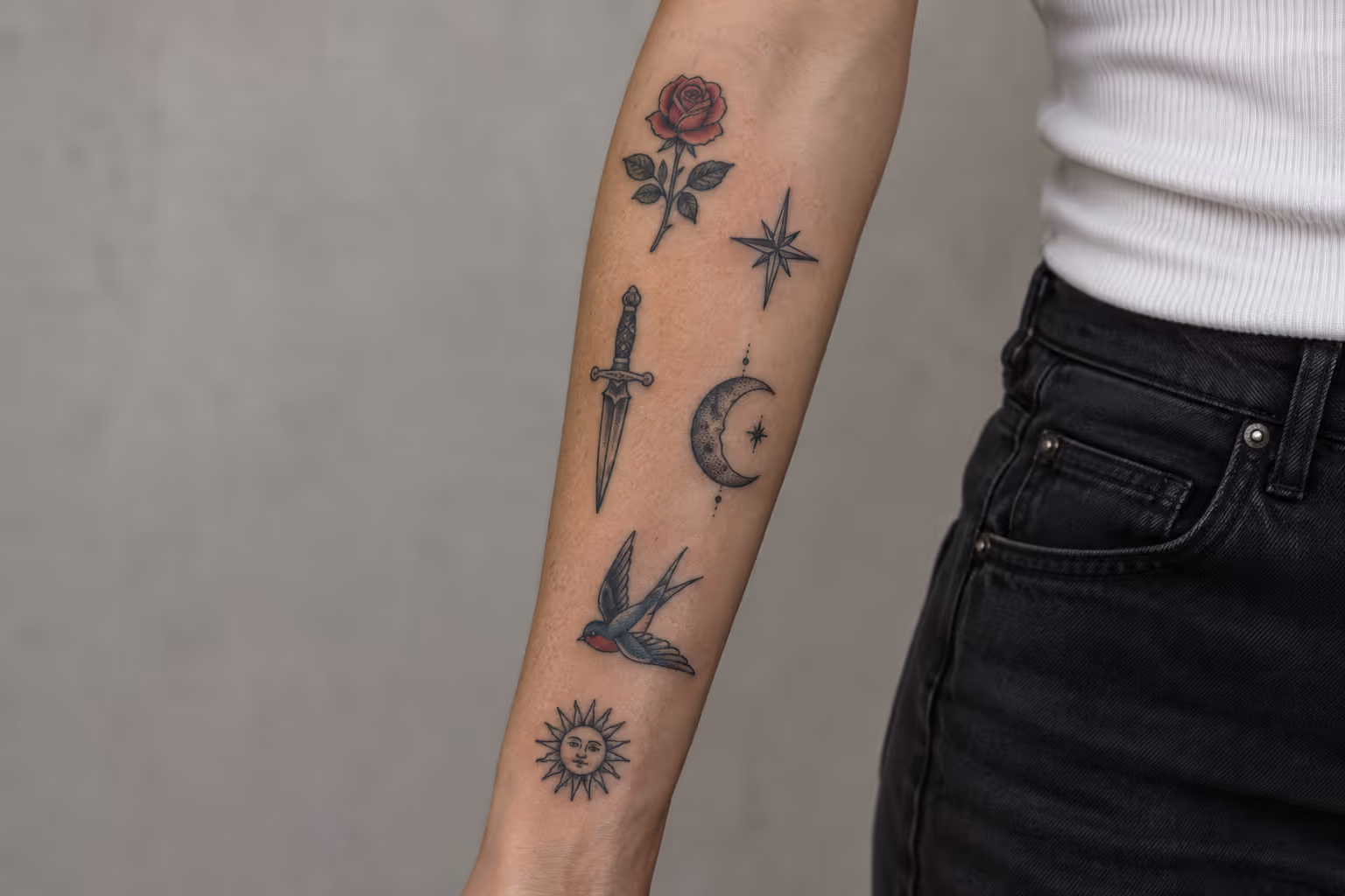 A forearm with small separate rose, star, dagger, moon, swallow and sun tattoos, bare skin between each piece.