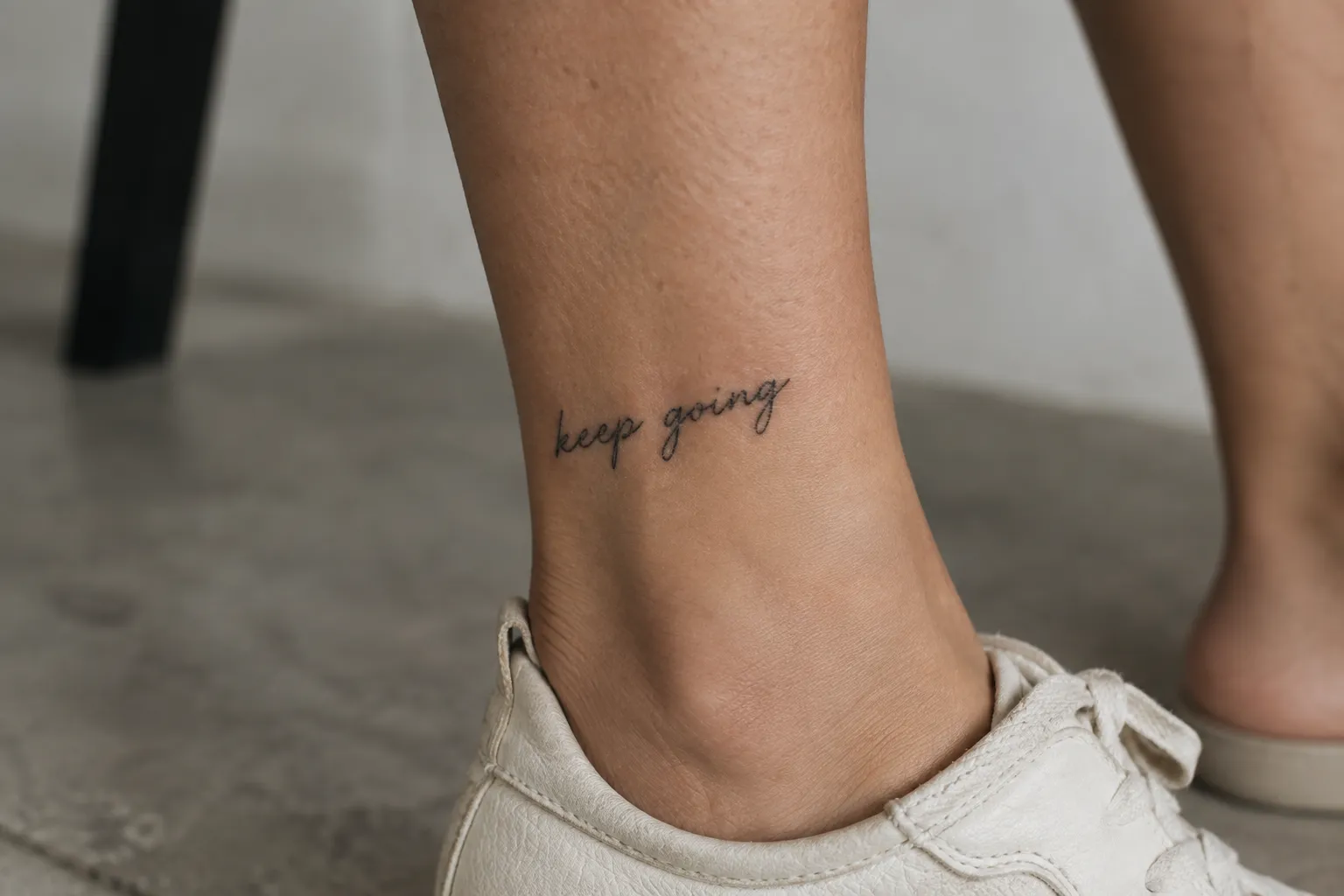 A person's ankle bears a small cursive 'keep going' tattoo above a white sneaker.