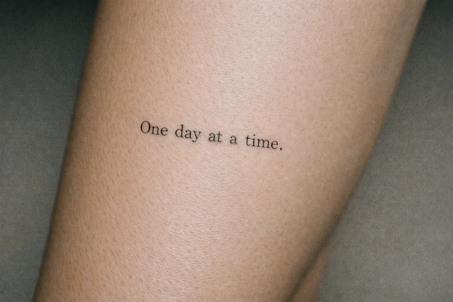 A small typewriter-style quote tattoo reading 'One day at a time.' on a person's lower leg.