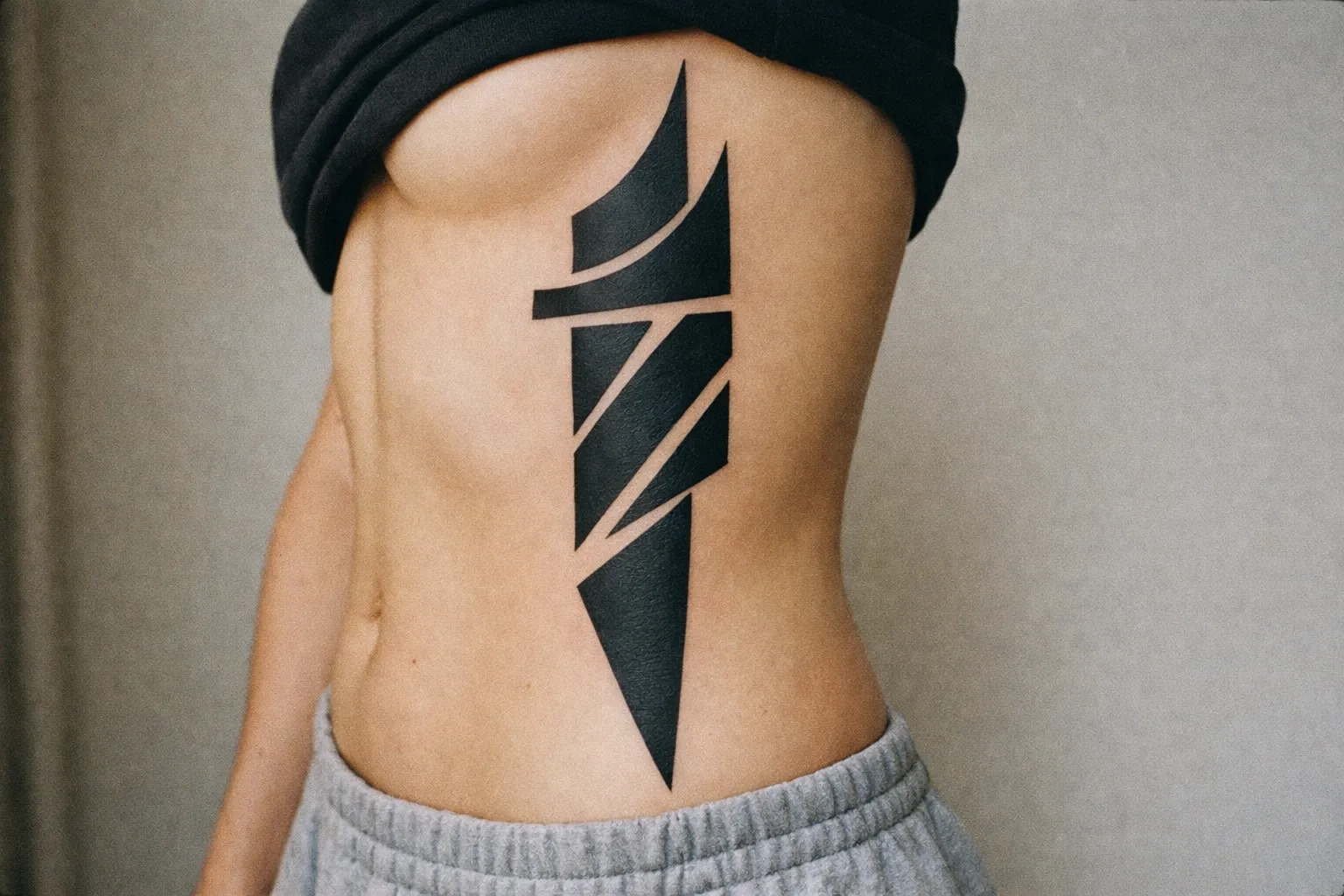 A person with a large geometric black tattoo on their ribcage stands indoors.