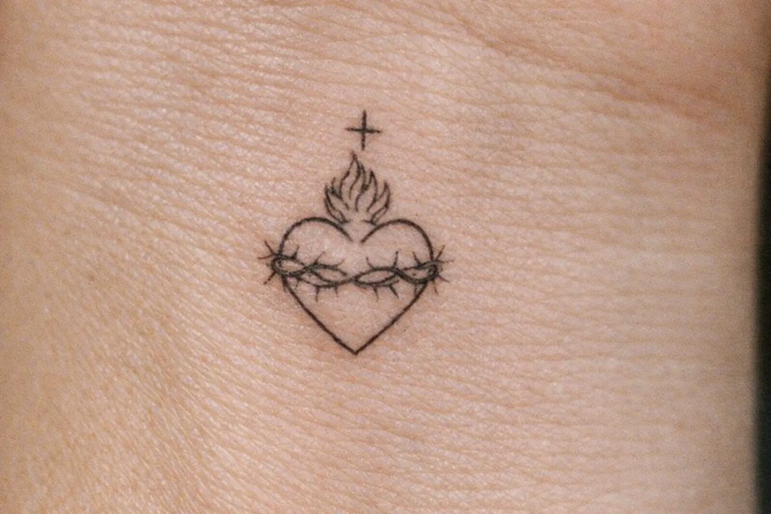 A small sacred heart tattoo on the skin features a heart with a crown of thorns and a cross above it.