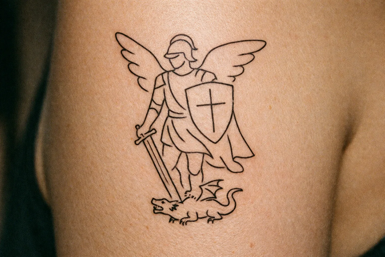 A Saint Michael tattoo depicting a winged figure with a sword and shield standing over a dragon on a person's arm.