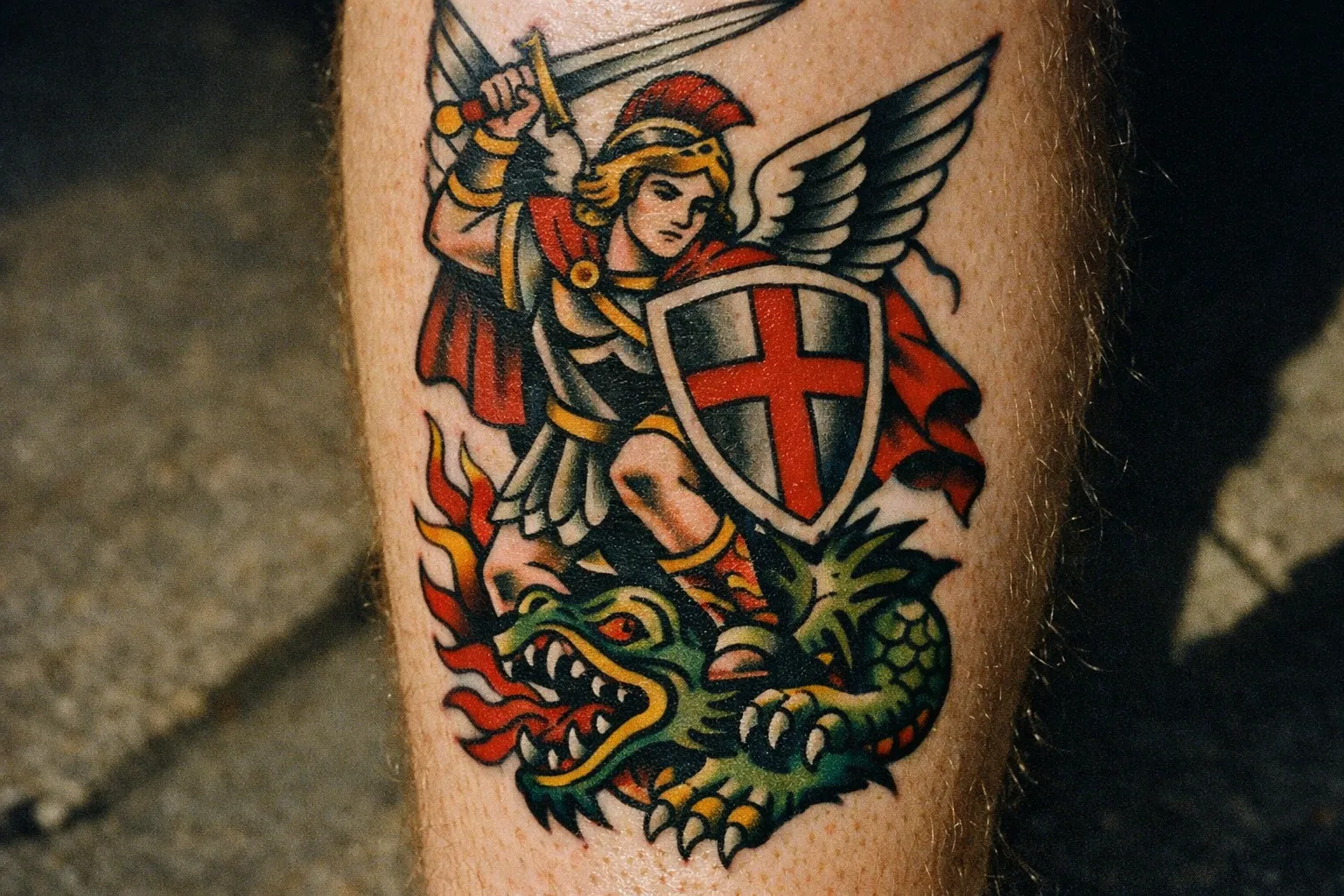 A tattoo of Saint Michael with a sword and shield standing over a dragon on a person's leg.
