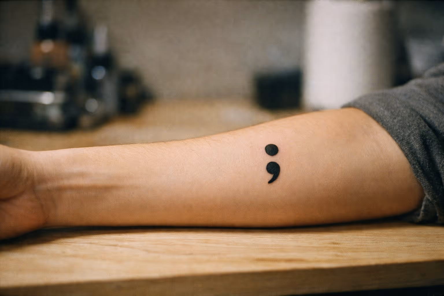 Editorial wide shot of a semicolon tattoo on a forearm, resting on a neutral surface with soft studio lighting and visible ink saturation.