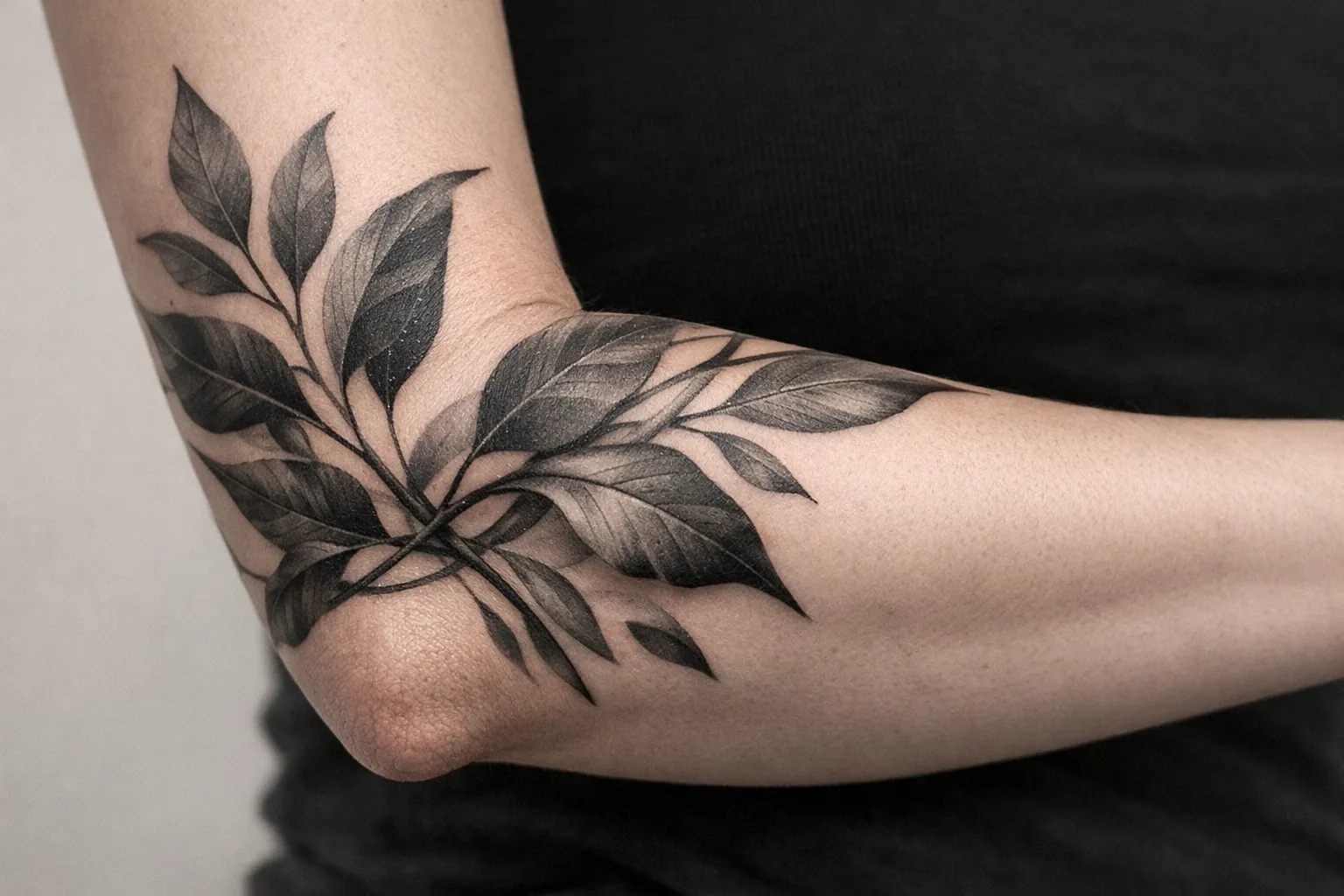 A person's forearm with a large black and gray leaf tattoo extending over the elbow.