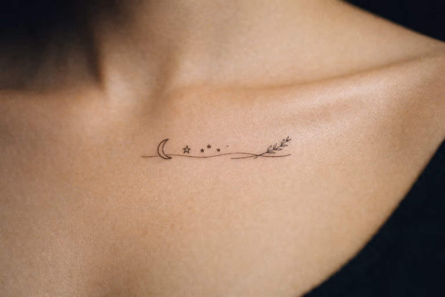 A small linework tattoo with a moon, stars, and branch on a person's collarbone.
