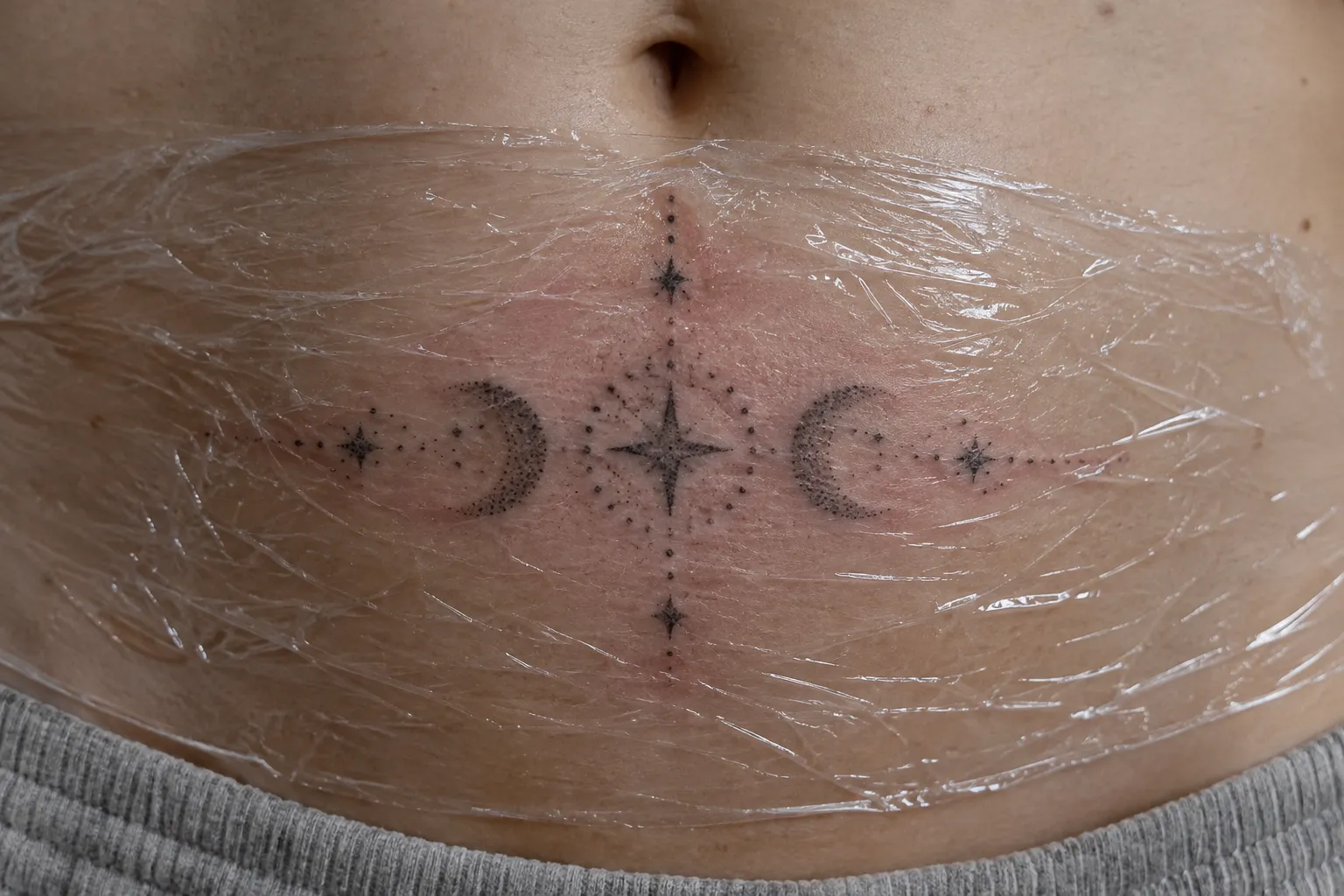 A fresh crescent moon and star tattoo sits across a person's lower abdomen beneath plastic wrap.