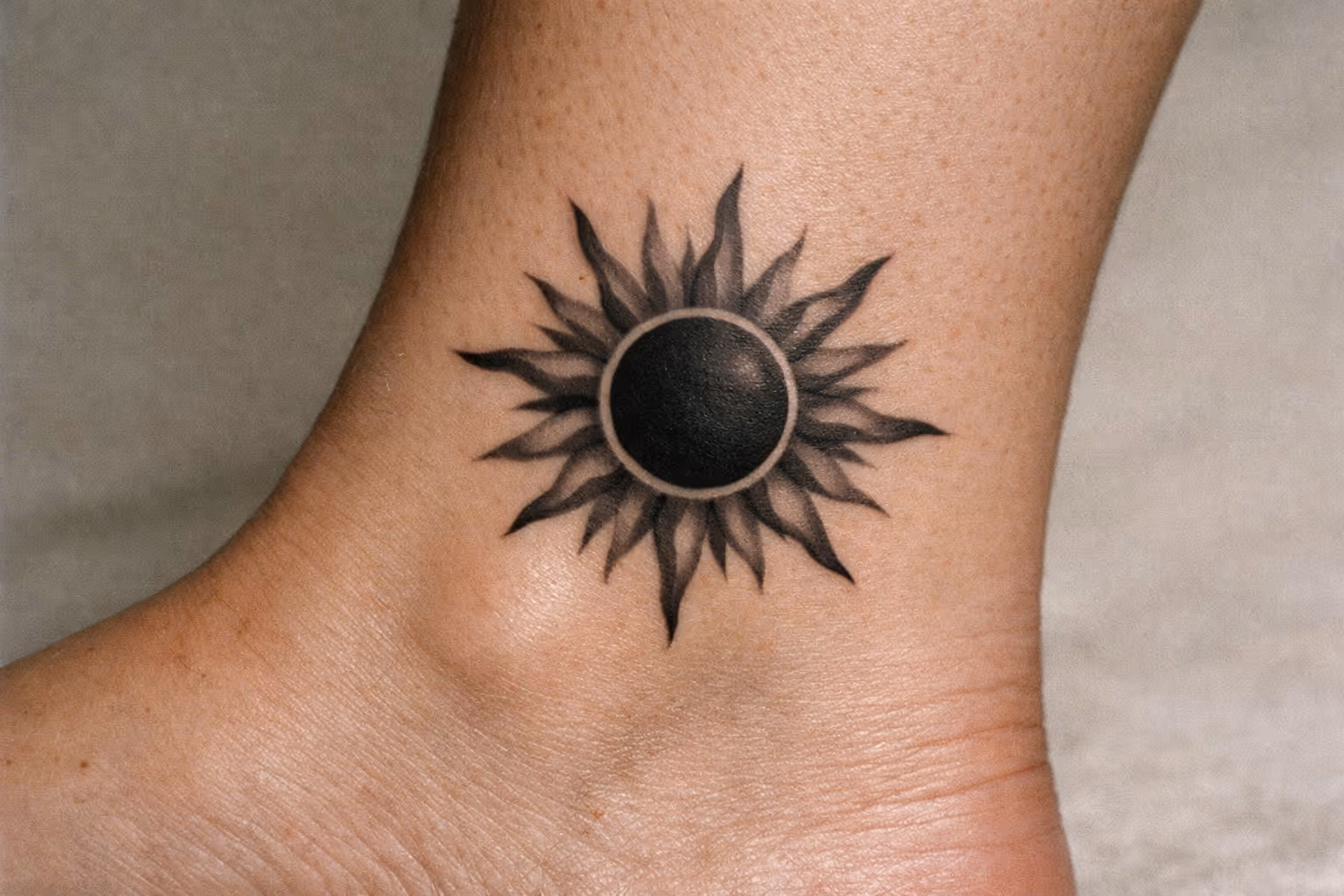 A black sun tattoo is inked on the side of a person's ankle.
