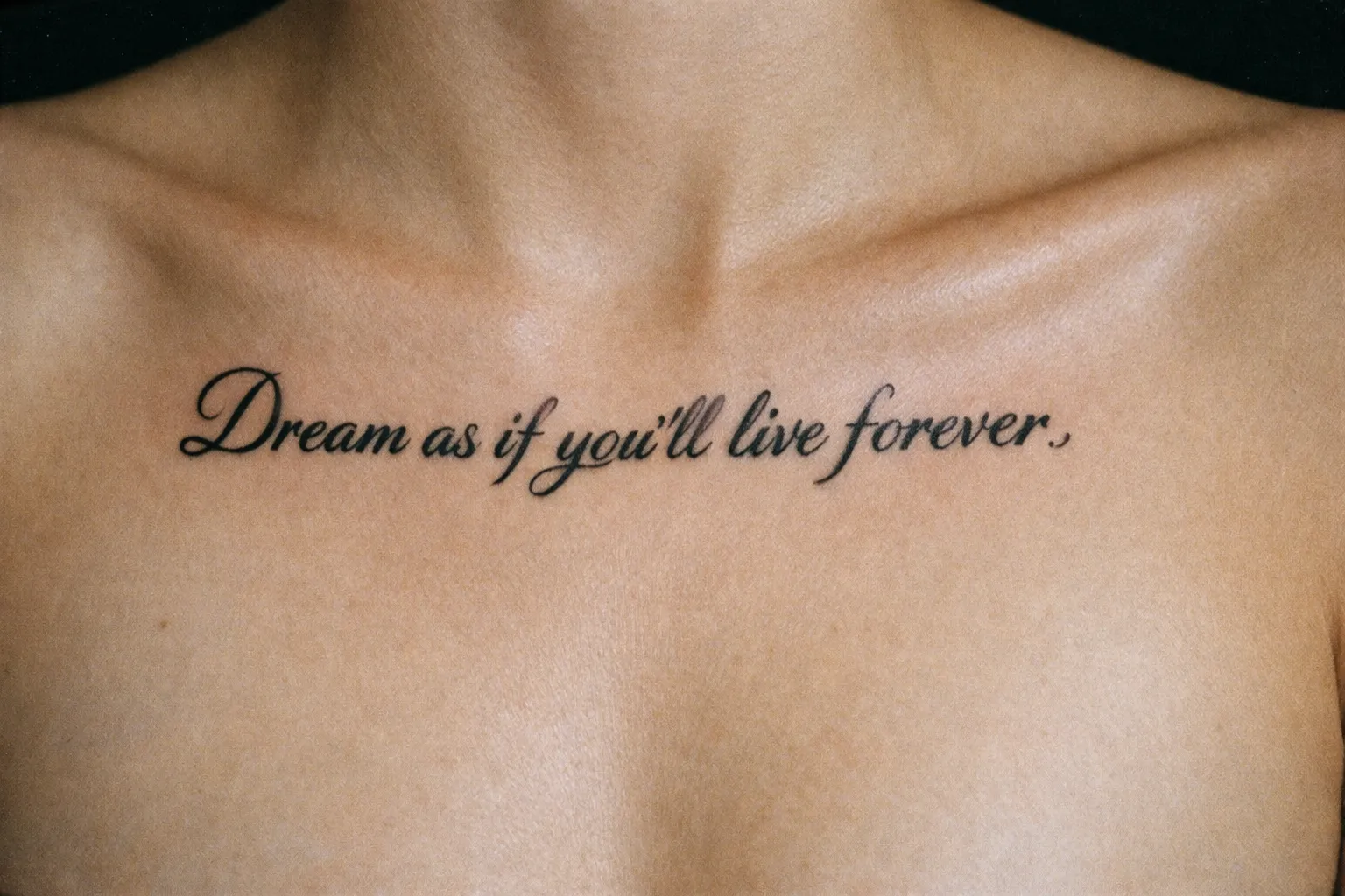 Script tattoo reading 'Dream as if you'll live forever.' on a person's upper chest.