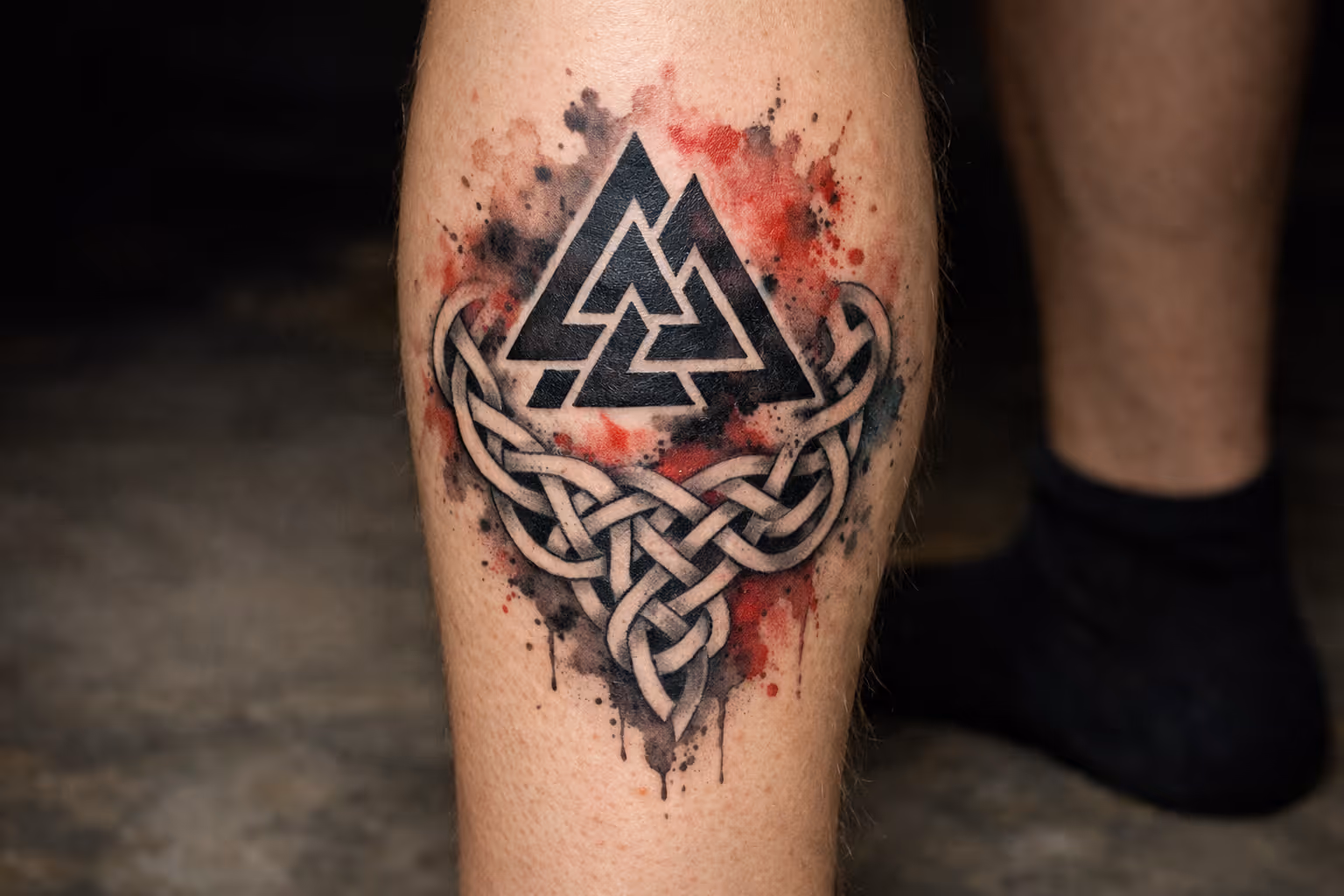 A close-up of a calf with a black Norse knotwork and Valknut tattoo against a blurred indoor floor.