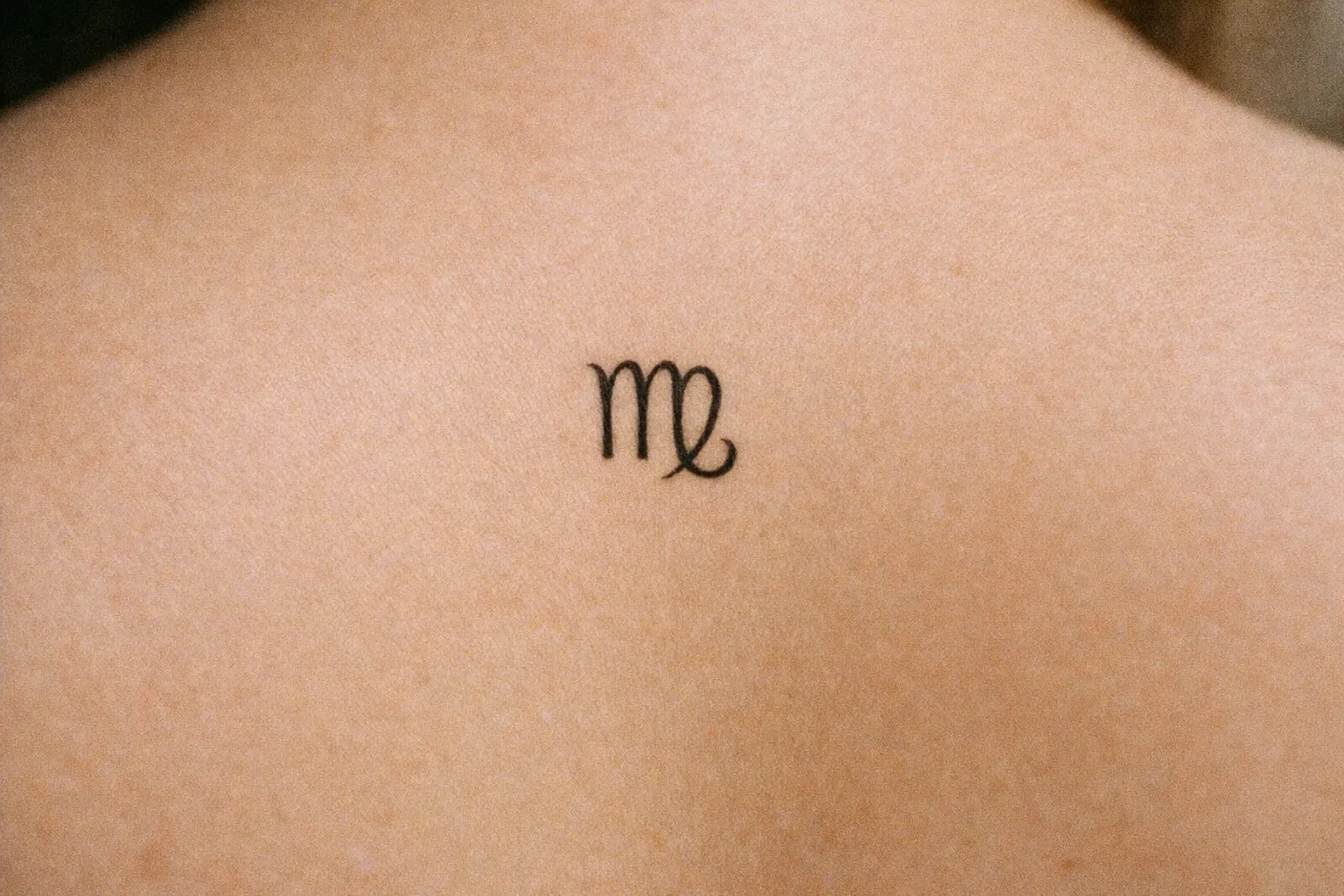 A small Virgo zodiac symbol tattoo on a person's upper back.