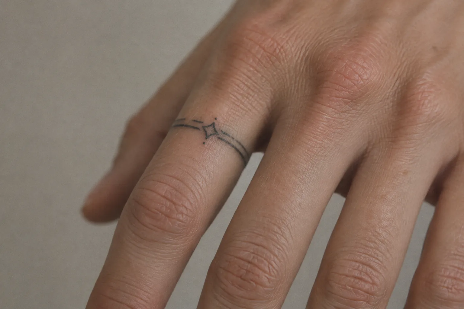 A hand with a delicate geometric ring tattoo on one finger against a plain background.