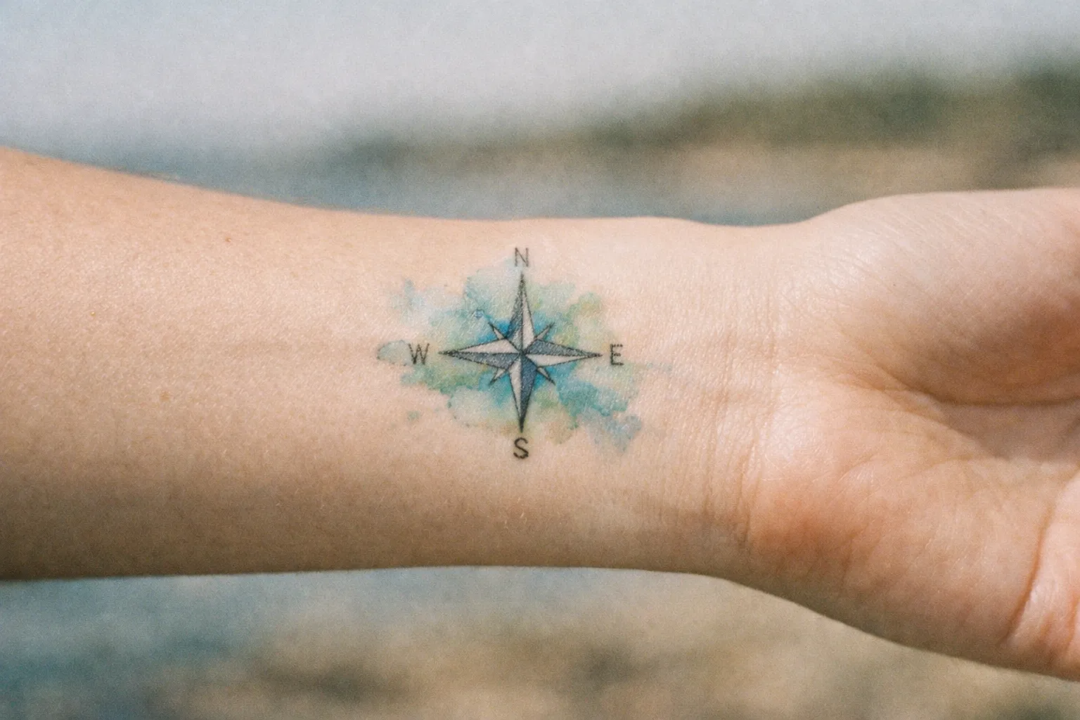 A compass rose tattoo with watercolor background is inked on the inside of a person's wrist.